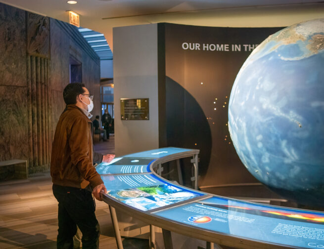 A person looking at a large blue, green, and brown rotating model of the Earth behind a curved table that is lit up with exhibit information.