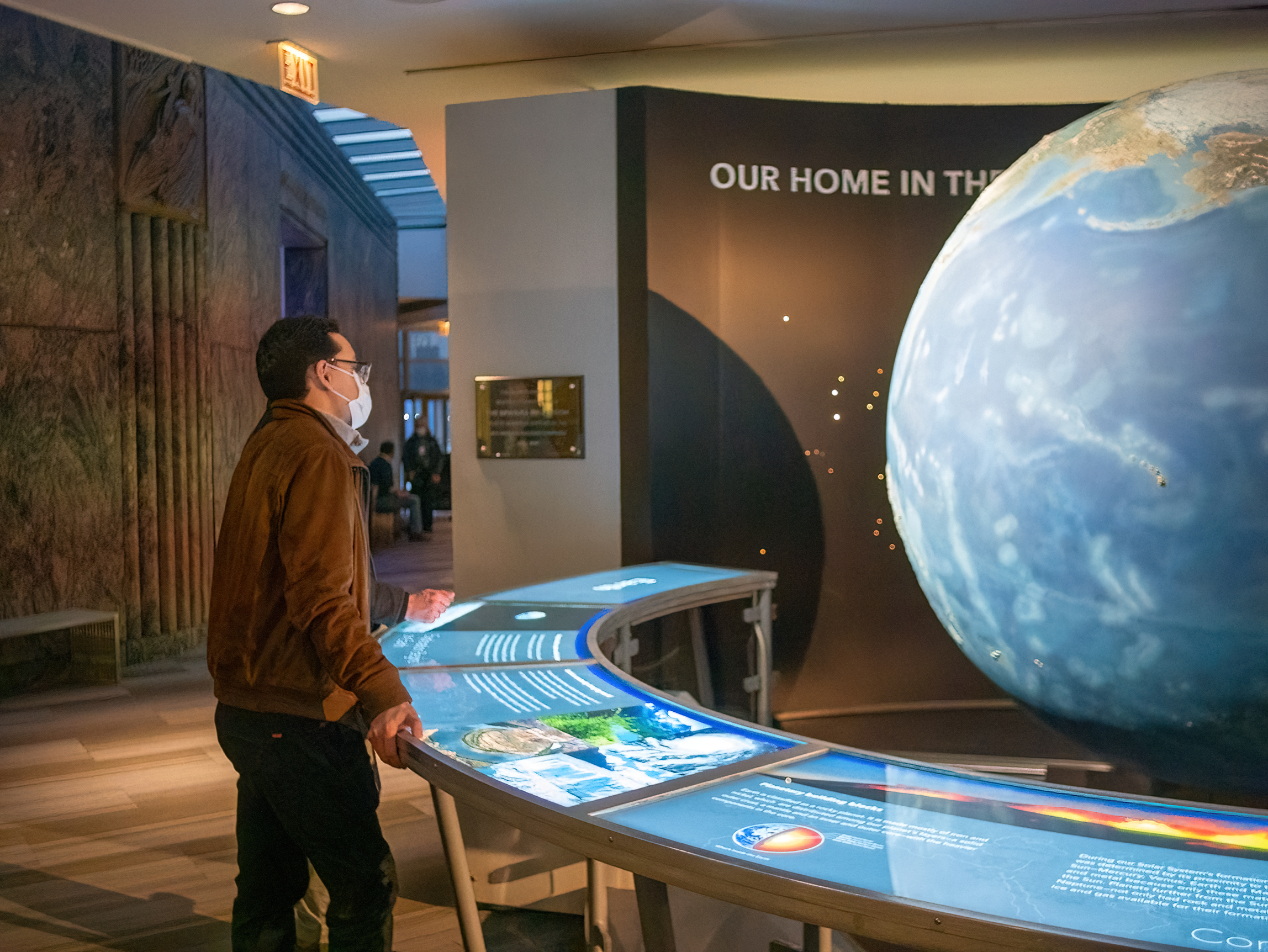 A person looking at a large blue, green, and brown rotating model of the Earth behind a curved table that is lit up with exhibit information.