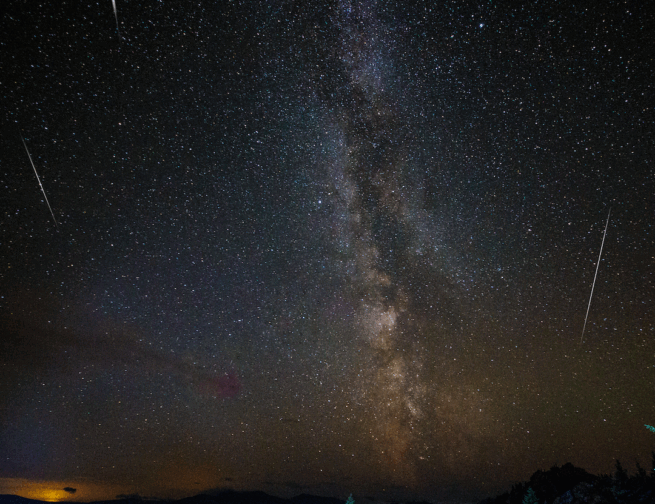 A dark starry night sky with three streaks of light across the sky. Deep hues of orange and purple from the Milky Way galaxy appear across the sky as well.