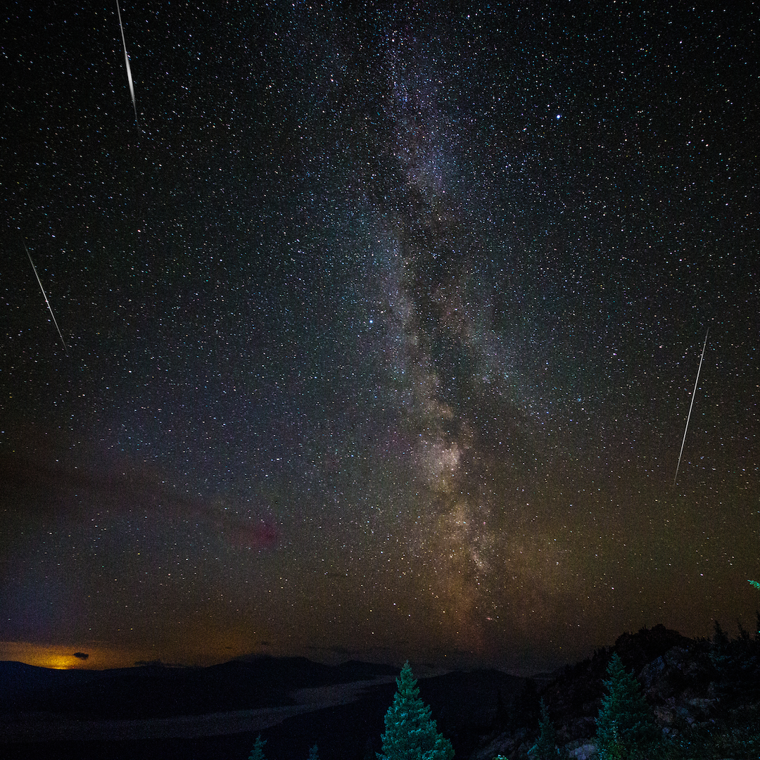A dark starry night sky with three streaks of light across the sky. Deep hues of orange and purple from the Milky Way galaxy appear across the sky as well.
