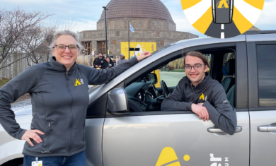 One person standing in front of a gray van with "Adler Planetarium" written on the van door and another person sitting inside the driver's seat of the van. In the background is the Adler Planetarium with a cloudy blue sky and a circular yellow logo with a gray car and "Astro Road Trip" written on it.