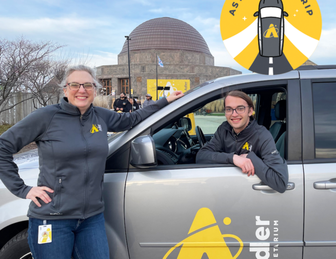 One person standing in front of a gray van with "Adler Planetarium" written on the van door and another person sitting inside the driver's seat of the van. In the background is the Adler Planetarium with a cloudy blue sky and a circular yellow logo with a gray car and "Astro Road Trip" written on it.