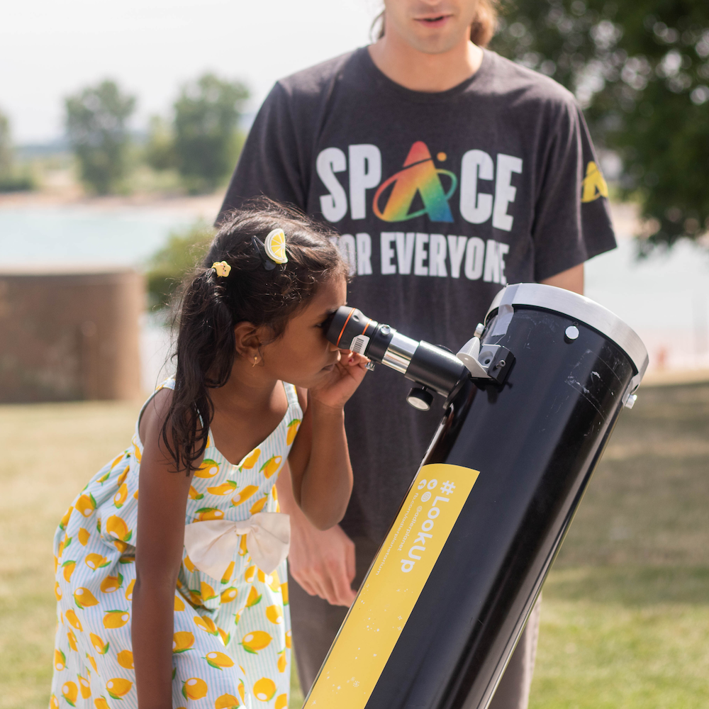 A child standing looking through a telescope during the day with a person behind the telescope wearing a shirt with "Space For Everyone"