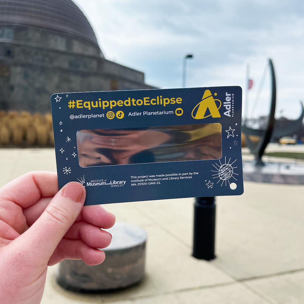 A hand holding a gray rectangular cardboard card with the center a black solar filtering film and white drawings of stars and the sun along the edges, hashtag "Equipped To Eclipse" and Adler Planetarium logo above the film, and Institute of Museum of Library Services logo and information below the film. In the background is a building and a large sundial. 