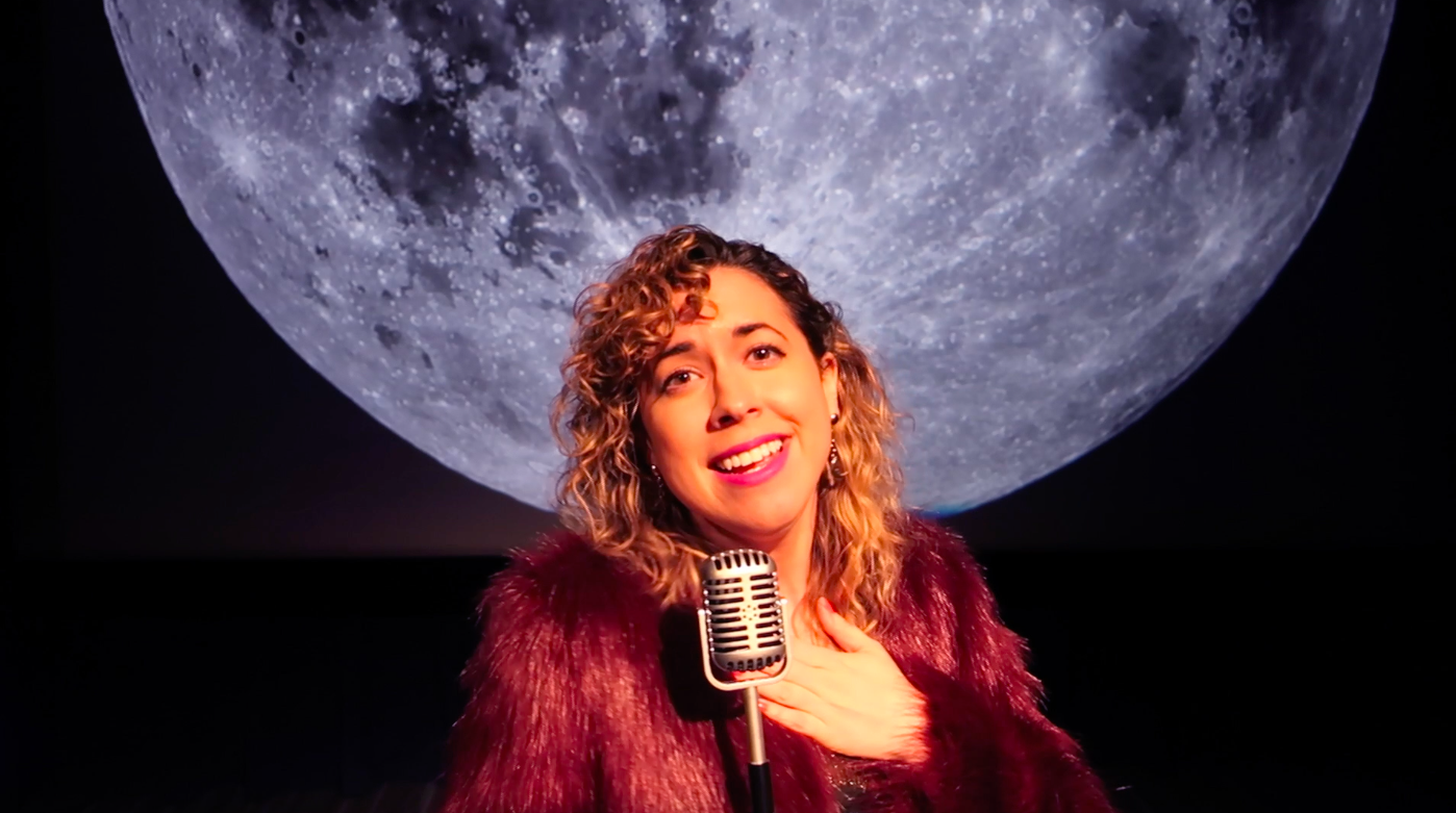 Meredith in a fuzzy purple coat singing into an old-school microphone with a Moon backdrop in the Johnson Star Theater