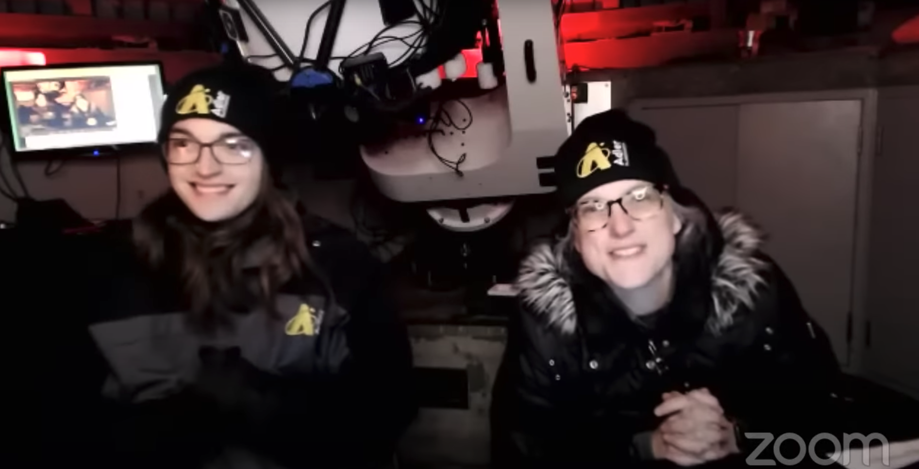 Astronomy educators Hunter and Michelle broadcast live from the Doane Observatory for a Sky Observers Hangout on Zoom. Seen behind Hunter and Michelle (who are wearing winter coats and hats), is a TV monitor, the Doane telescope, and a red light.