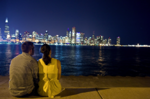 Couple sitting on the ground in front of Lake Michigan, viewing the Chicago skyline in the night sky. 