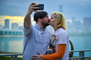 A couple standing outside of the Adler Planetarium, taking a selfie of them kissing in front of Lake Michigan and the Chicago Skyline.