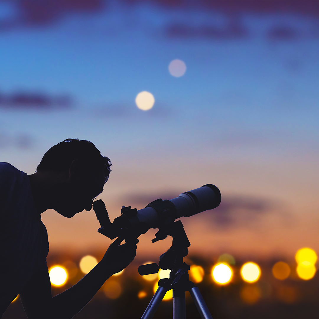Silhouette of a person looking through a small telescope on a tripod at dusk