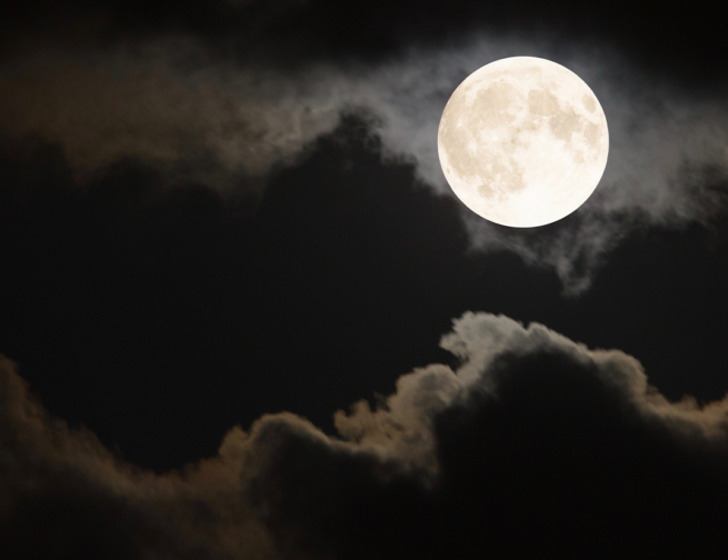 The full Moon shines bright white in a dark, cloudy sky.