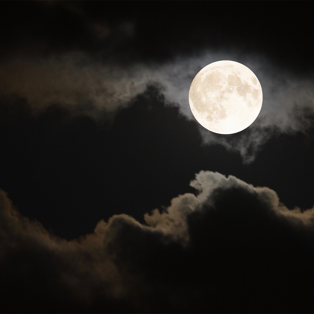 The full Moon shines bright white in a dark, cloudy sky.