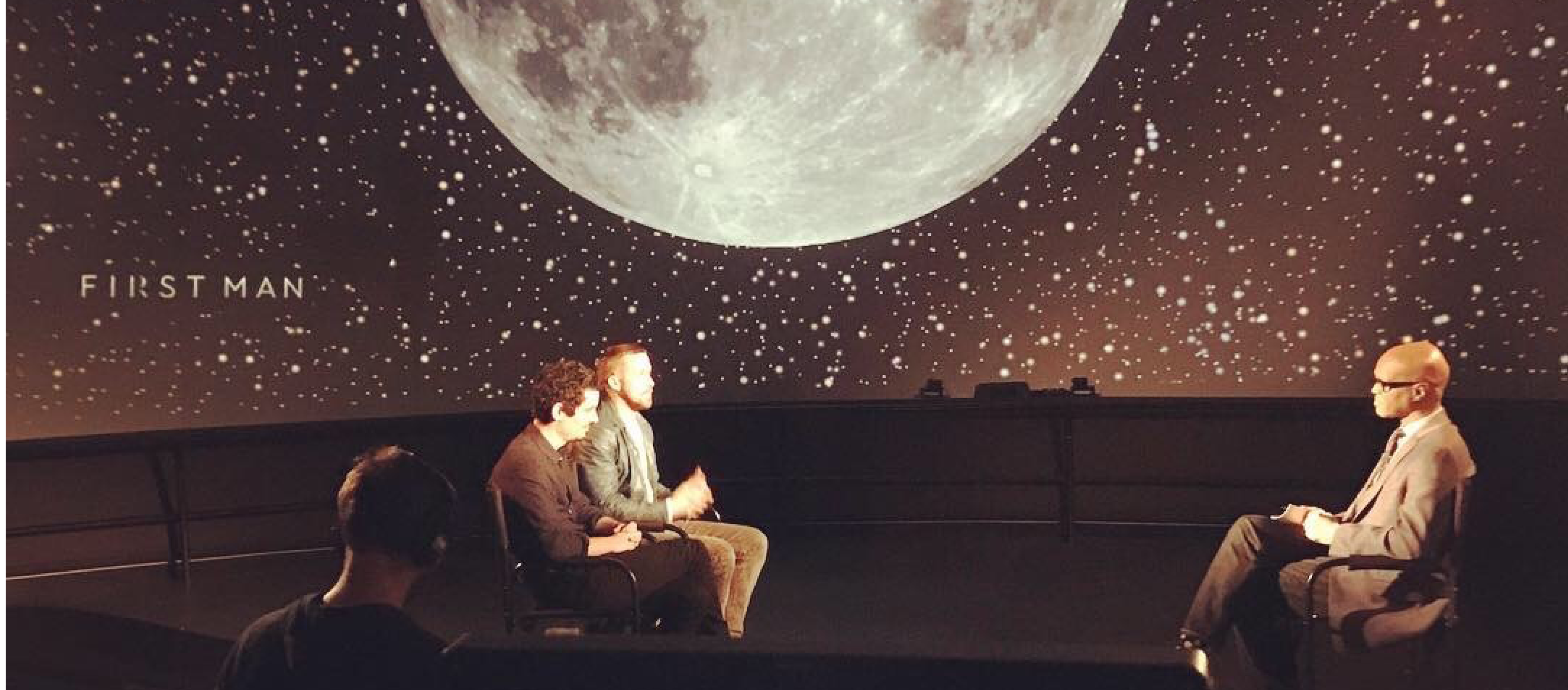 Grainger Sky Theater with Moon and stars projected onto the dome. Middle ground is award-winning actor, Ryan Gosling, and award-winning director, Damien Chazelle sitting across from a presenting african american male, bald with glasses, being interviewed by a reporter for the press junket of First Man in October 2018. Sound assistant can be seen in the foreground.