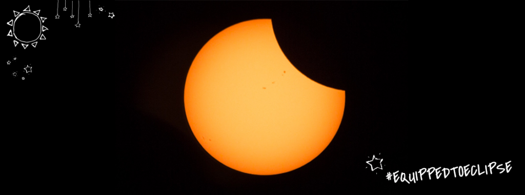 The Sun with about 1/4 of the upper right part of its yellow and orange disk partially eclipsed by the Moon and white drawings of the sun and stars around the outer edges of the image with the hashtag "Equipped To Eclipse"