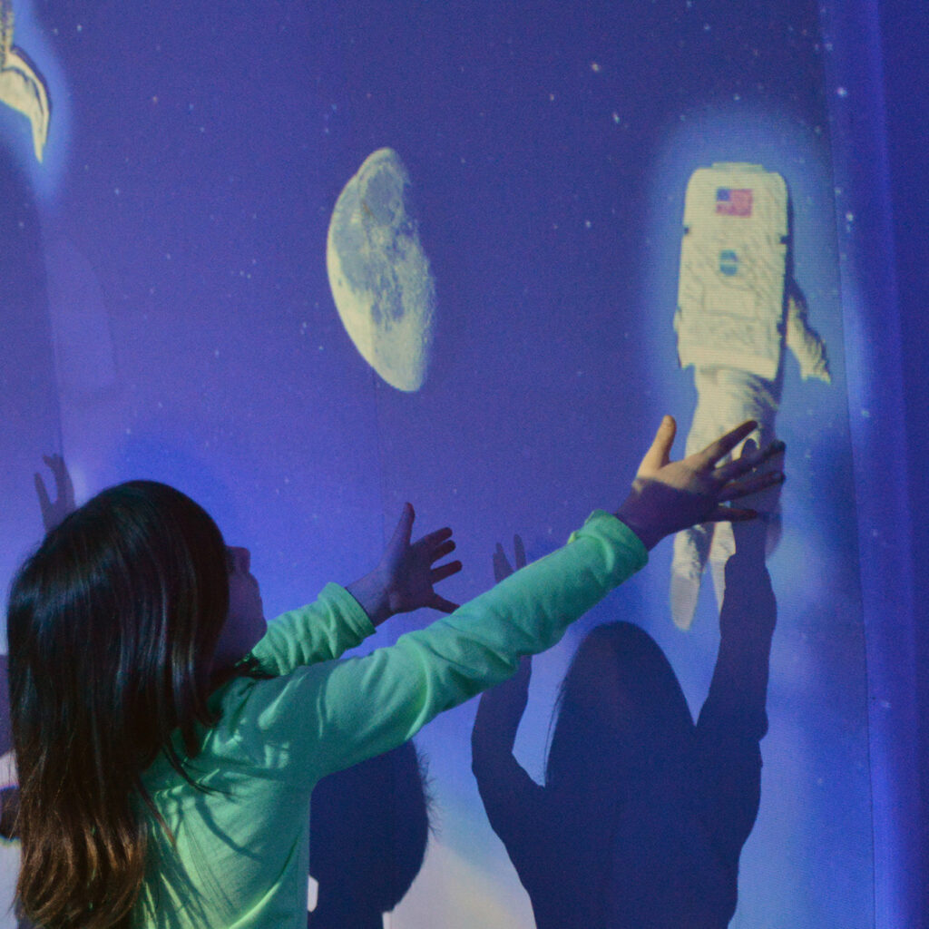 A kid in a green shirt reaching up to a wall showing a projected astronaut graphic.