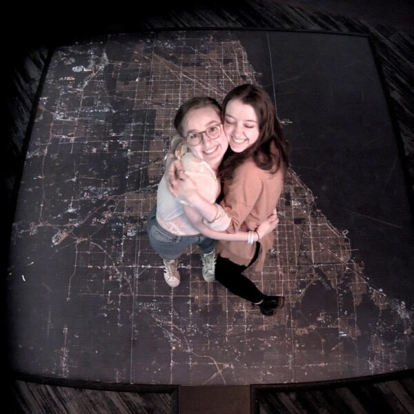 Two women embrace while standing on an aerial image of Chicago at night