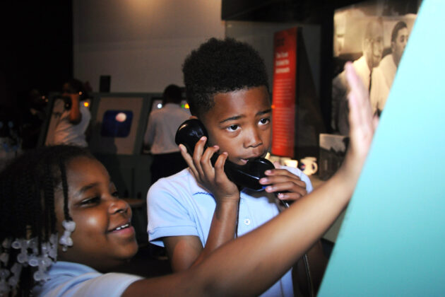 A boy holds a phone receiver to his ear while a girl pushes buttons on an installation