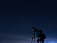 A silhouette of a person kneeling in front of a telescope, aimed at the night sky. The background shows a dark blue night sky with dim stars in the distance.