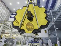 The JWST is in the center with its yellow hexagonal mirrors facing the front as a team of people and a crane lifts up the telescope in a room.