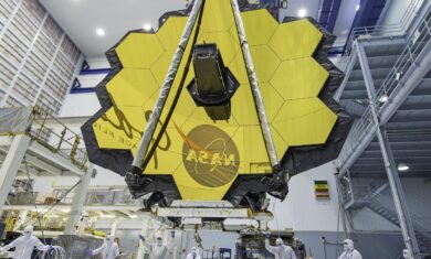 The JWST is in the center with its yellow hexagonal mirrors facing the front as a team of people and a crane lifts up the telescope in a room.