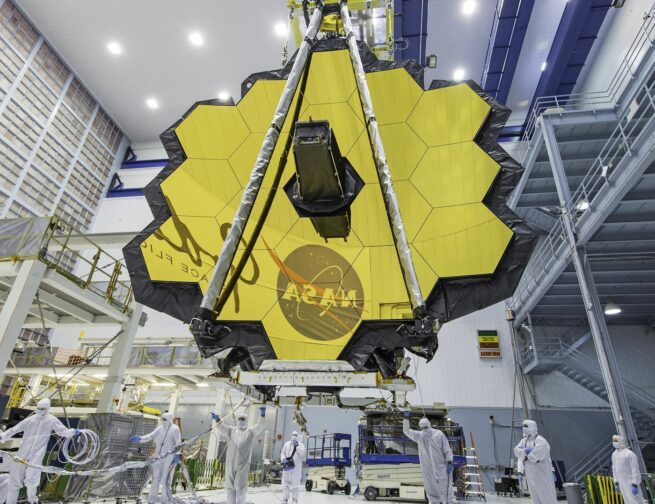 The JWST is in the center with its yellow hexagonal mirrors facing the front as a team of people and a crane lifts up the telescope in a room.