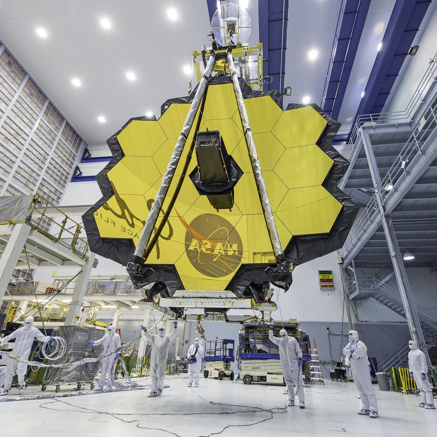 The JWST is in the center with its yellow hexagonal mirrors facing the front as a team of people and a crane lifts up the telescope in a room.