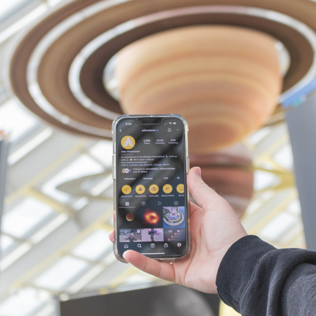 A hand holding up a cell phone showing the Adler Planetarium's Instagram page