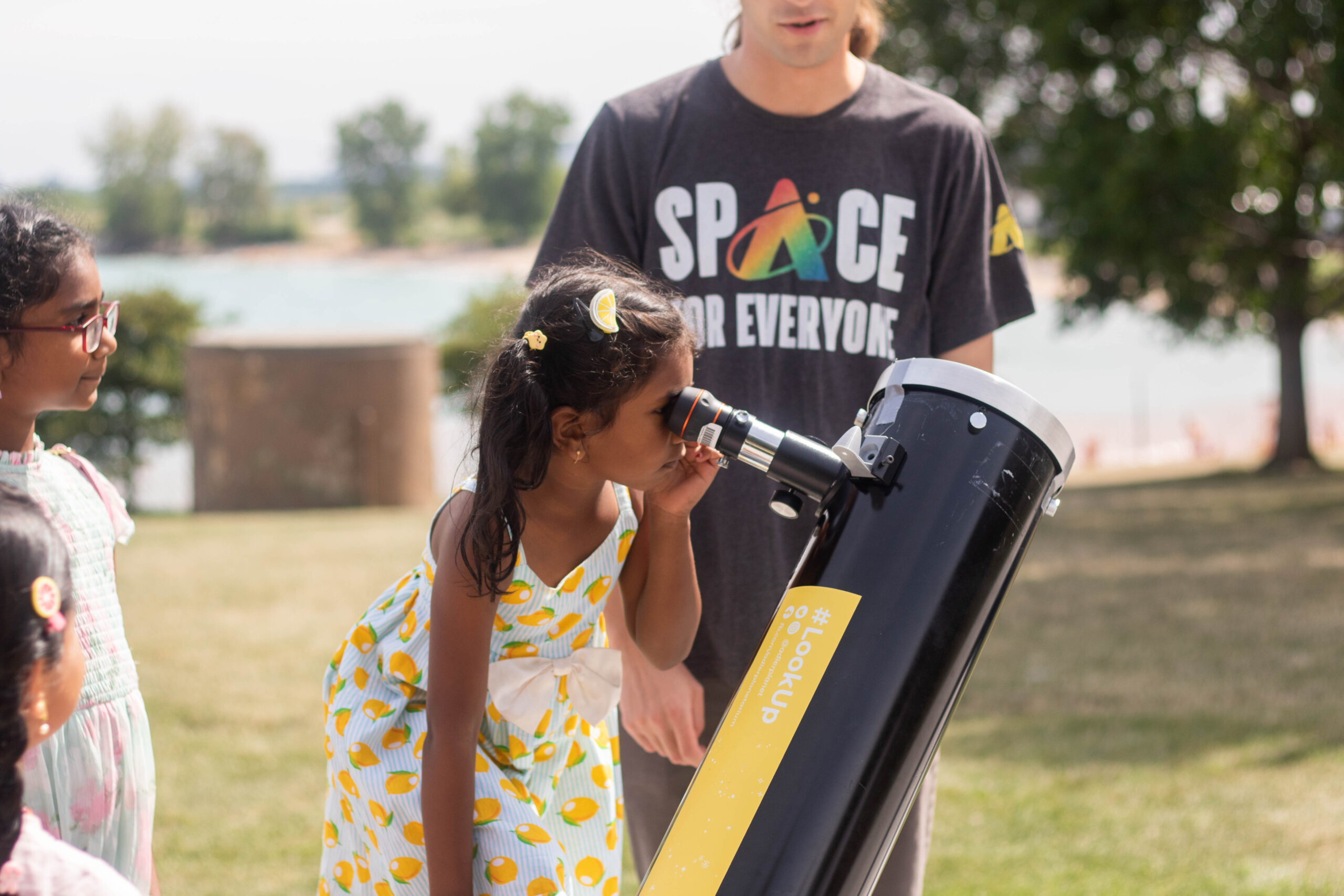 Public observing educator with Space for Everyone t-shirt standing by as a child with a lemon dress looks through a telescope on the observing terrace.