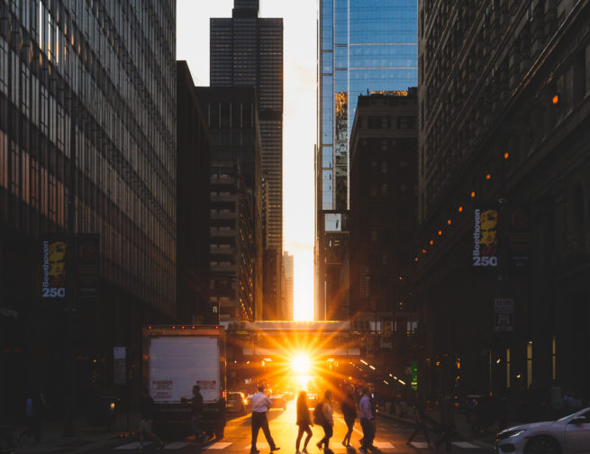 Pedestrians walking (during Chicagohenge) across a crosswalk while the Sun sets in the distance with the Willis Tower in view.
