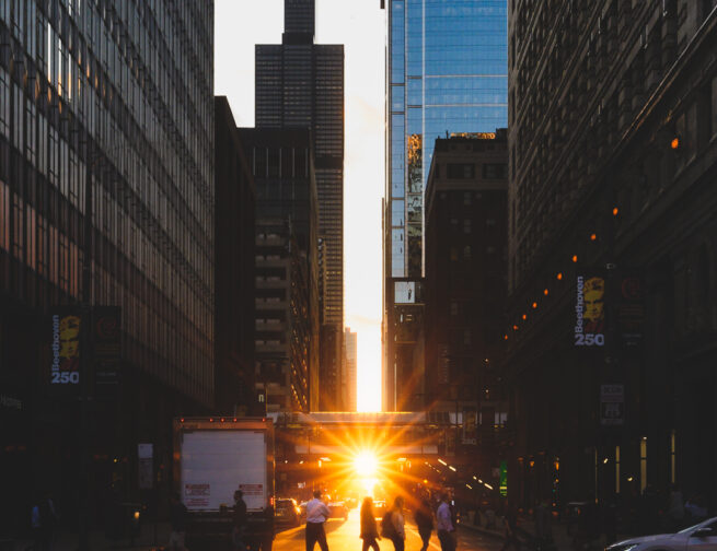 Pedestrians walking (during Chicagohenge) across a crosswalk while the Sun sets in the distance with the Willis Tower in view.