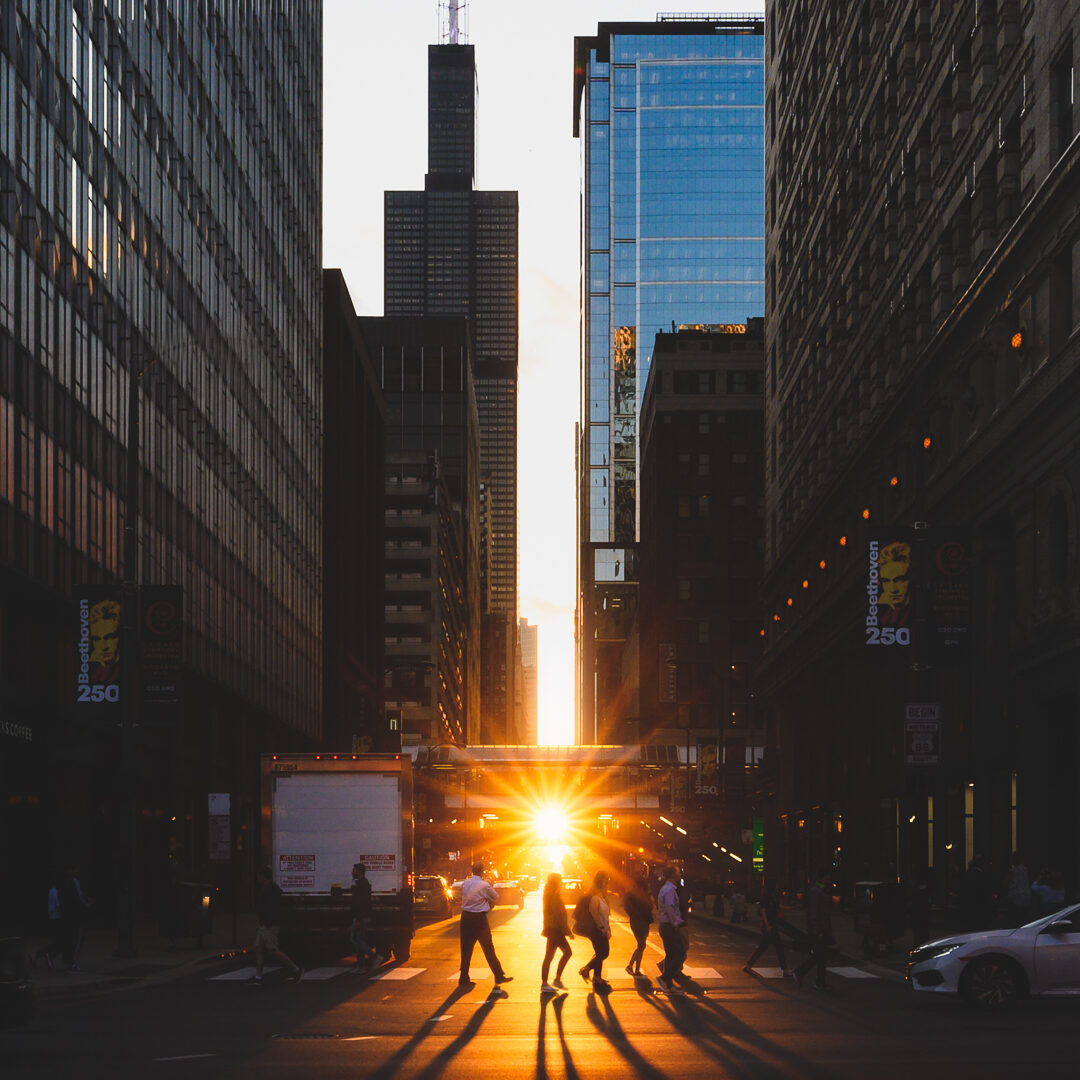 Pedestrians walking (during Chicagohenge) across a crosswalk while the Sun sets in the distance with the Willis Tower in view.